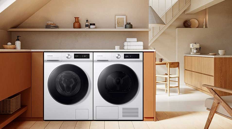 A laundry room features a white Samsung washer and dryer set under a shelf with decor, set in a modern, beige-toned interior with wood accents.