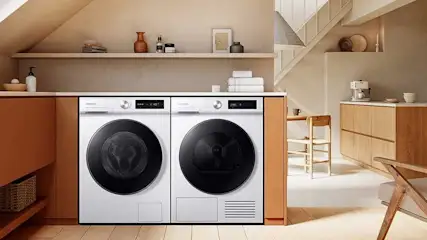 A laundry room features a white Samsung washer and dryer set under a shelf with decor, set in a modern, beige-toned interior with wood accents.