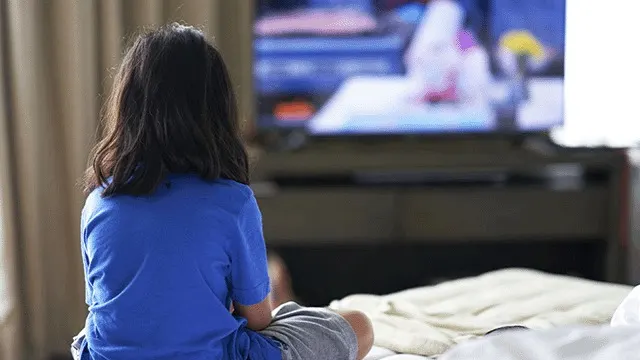 Een persoon zit op een bed, gekleed in een blauw shirt en grijze broek, kijkend naar een tv. Gordijnen op de achtergrond.