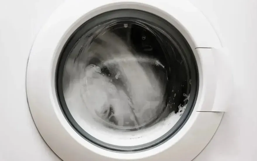 Een witte wasmachine draait, schuim en water zijn zichtbaar achter het ronde glazen deurvenster. Er is geen merknaam te zien op de machine.
