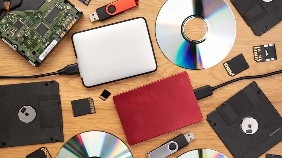 Divers supports de stockage numériques variés : disques durs, clés USB, cartes SD et disquettes sur fond bois clair.