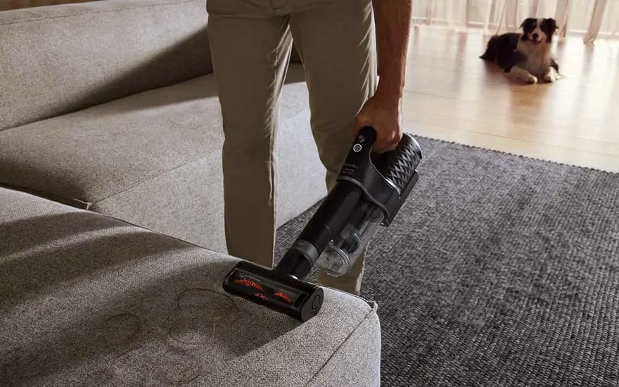 Iemand stofzuigt een grijze bank met een kruimeldief op een grijze vloerkleed. Een hond ligt op een houten vloer.