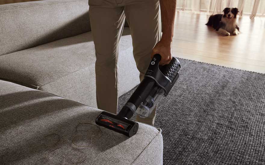 Une personne utilise un aspirateur noir sur un canapé gris. Un tapis gris se trouve à côté. Un chien est sur le plancher.