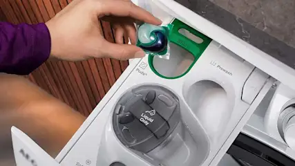 Een hand plaatst een wasmiddelpod in een witte wasmachine-lade met compartimenten voor pods, voorwas en vloeibaar wasmiddel.