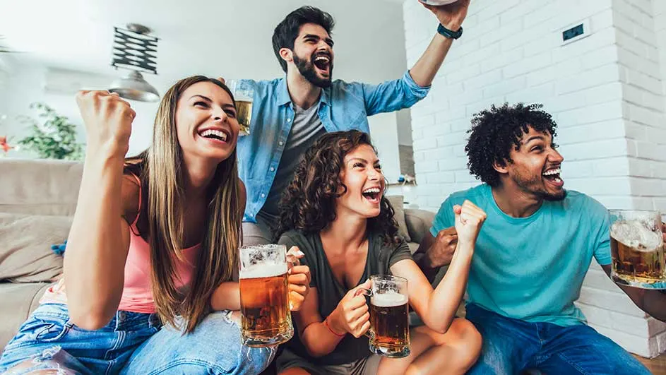 Un groupe tient des verres de bière. Le mur est blanc, fait de briques. Une lumière est accrochée au plafond.