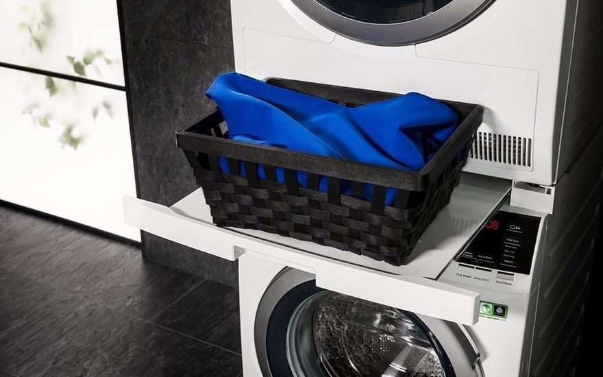 Stacked washer and dryer with a blue cloth in a basket. Black tiled floor and a window in the background.