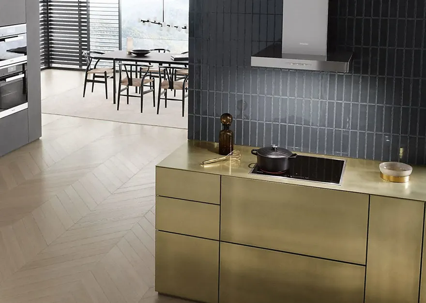 Cuisine moderne avec comptoir doré, plaque de cuisson noire, et crédence bleu-gris. Table et chaises visibles en arrière-plan.