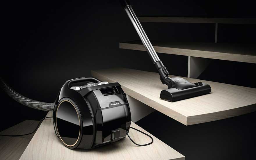 Un aspirateur noir avec un tuyau gris est posé sur une marche d'escalier beige. La brosse d'aspirateur est sur la marche suivante.