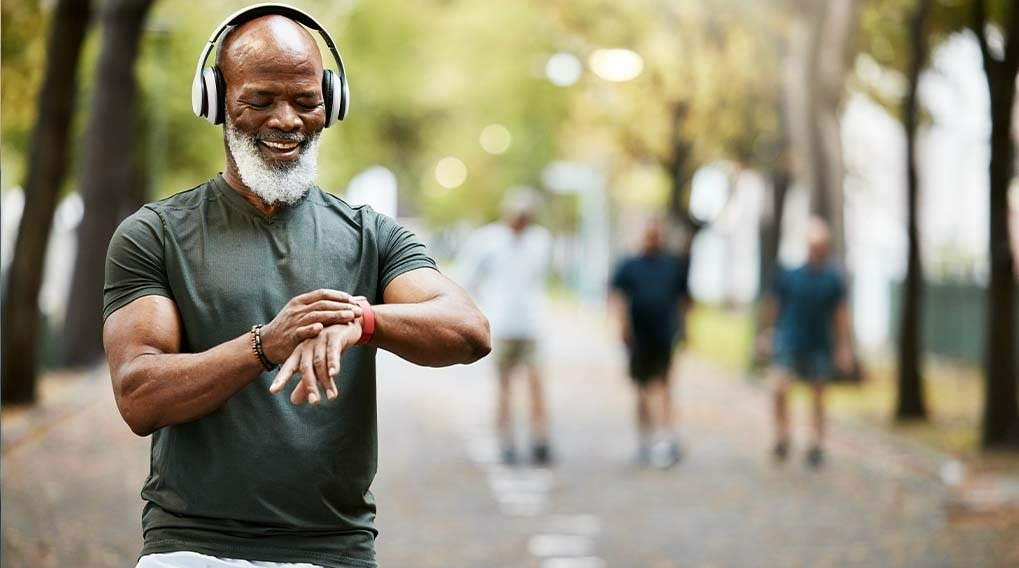 Homme avec un casque audio regardant une montre connectée, sur un chemin bordé d'arbres et d'autres personnes.