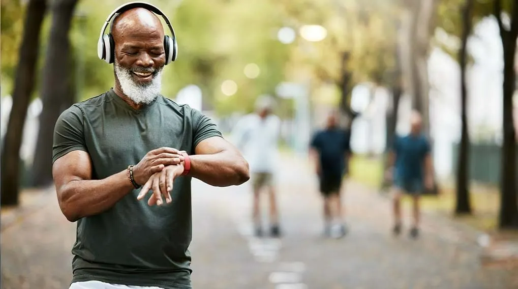 Homme avec un casque audio regardant une montre connectée, sur un chemin bordé d'arbres et d'autres personnes.