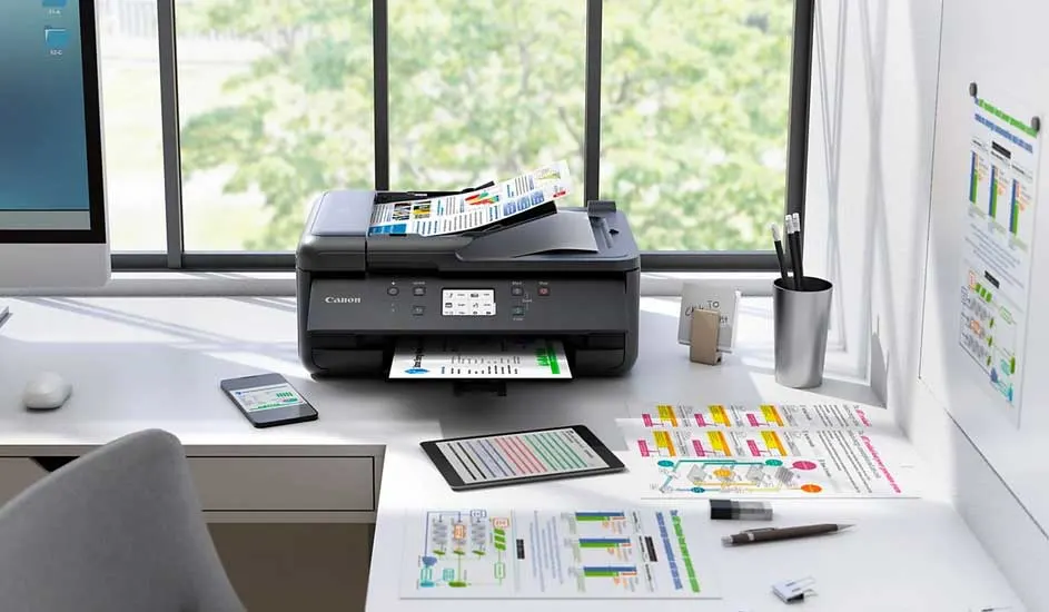 Een printer op een bureau met een telefoon, tablet, papier en een computer tegen een raam.