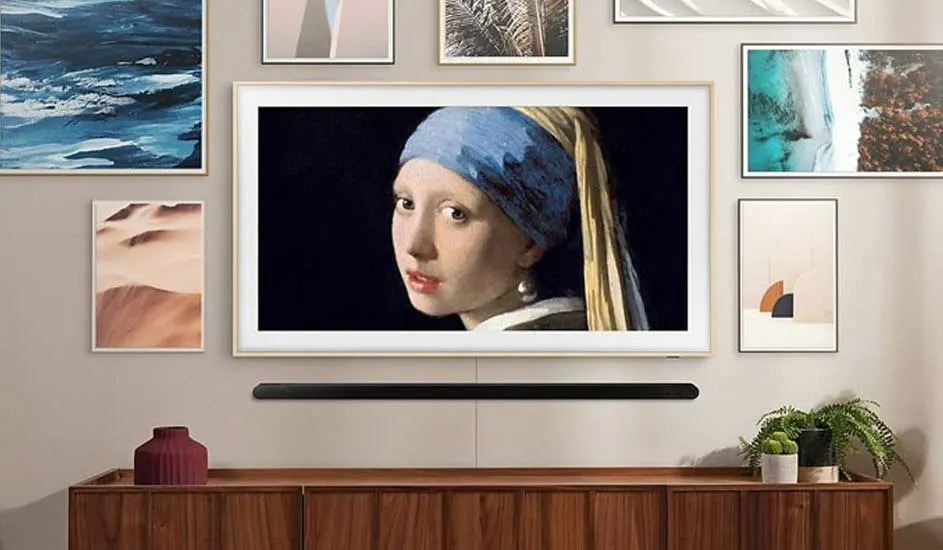 Een Samsung The Frame tv hangt aan de muur tussen schilderijen en toont een beroemd kunstwerk, met een soundbar eronder en planten op het meubel.
