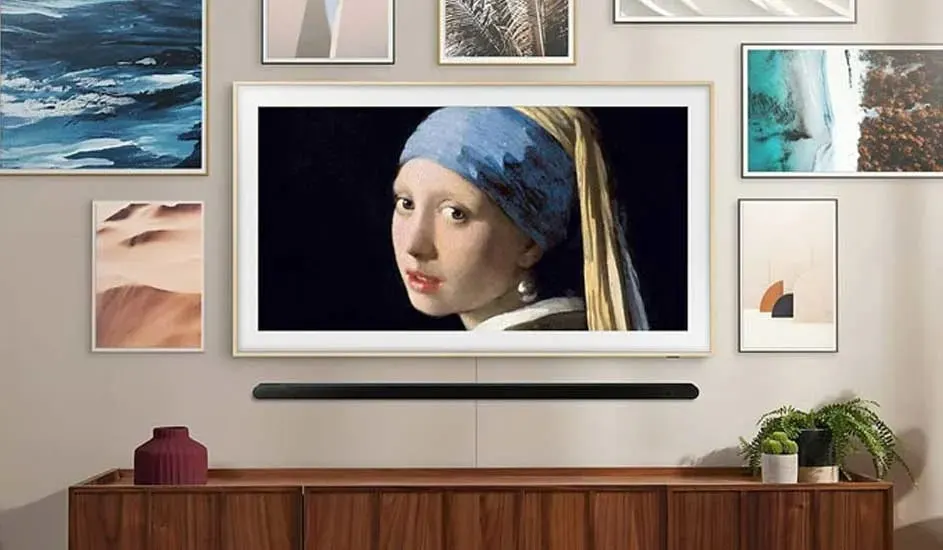 Une télévision Samsung The Frame est accrochée au mur parmi des tableaux et affiche une œuvre d’art célèbre, avec une barre de son en dessous et des plantes sur le meuble.