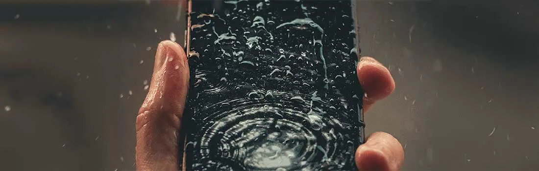 Un téléphone noir est tenu, couvert de gouttes d'eau, créant des ondulations concentriques sur l'écran.