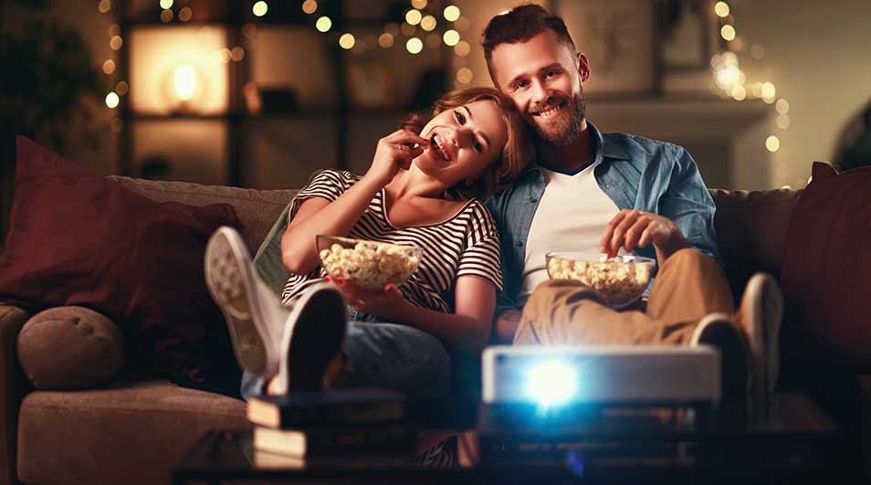 Living room scene with a projector beaming light. Couch, pillows, and popcorn bowls visible. Background has lights.