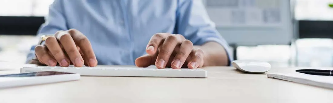 Gros plan sur un clavier blanc avec des mains, une souris, un bloc-notes et un stylo noir sur un bureau clair.