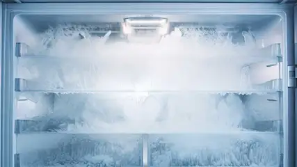 Intérieur d'un réfrigérateur blanc vide, avec plusieurs étagères et une forte accumulation de givre. Une lumière est allumée en haut.