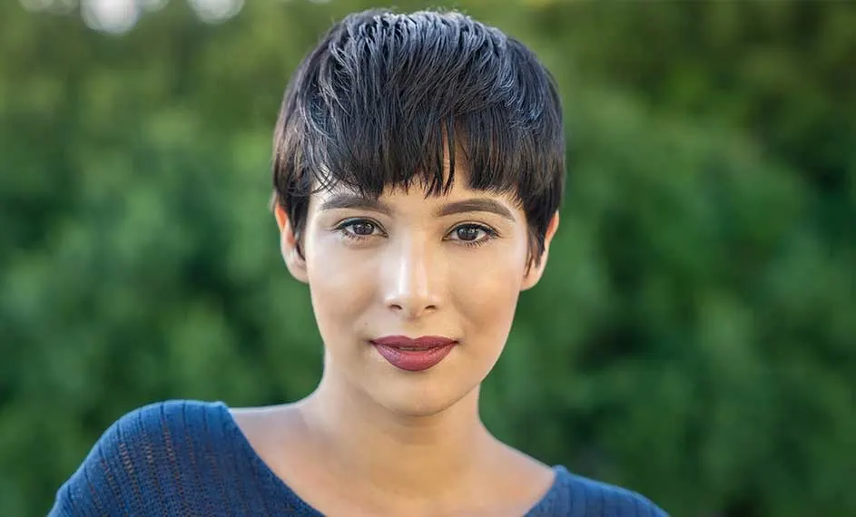 Een close-up toont een persoon met kort, donker haar en een blauwe trui, tegen een groene achtergrond.