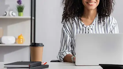 Bureau blanc avec ordinateur portable, café et carnets noirs. Étagère avec des plantes et des objets décoratifs en arrière-plan.