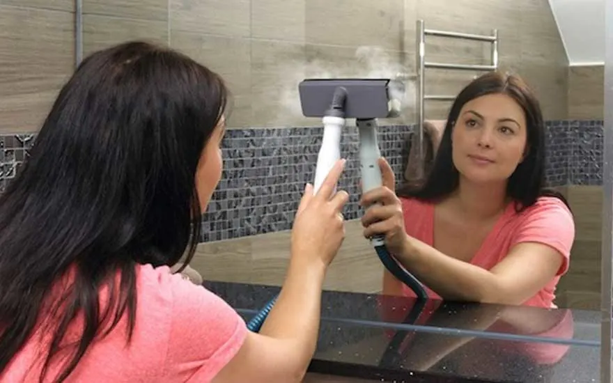 Une personne utilise un nettoyeur vapeur gris et blanc sur un miroir dans une salle de bain avec des carreaux décoratifs.