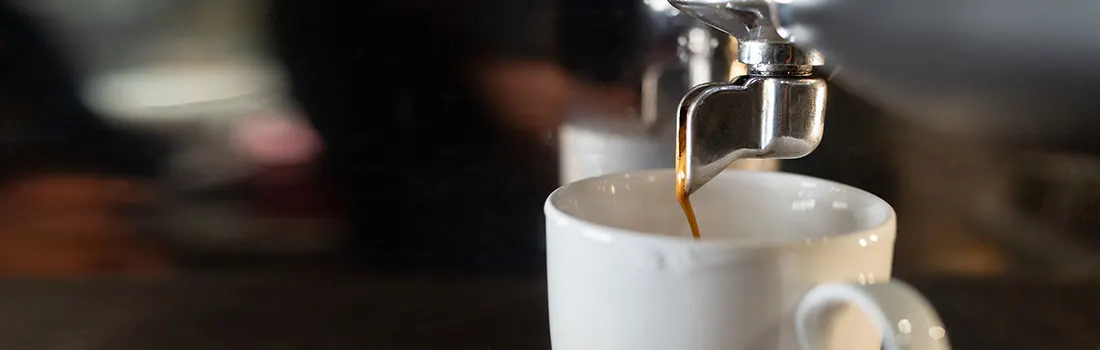 Une tasse blanche est remplie de café provenant d'une machine à expresso en métal sur un fond sombre.