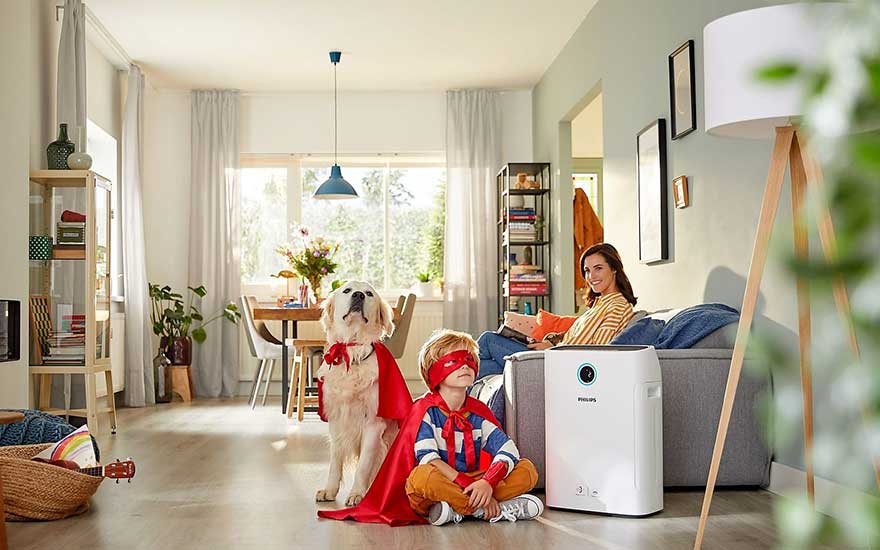 Intérieur lumineux avec purificateur d'air blanc Philips, chien et enfant portant des capes rouges. Table à manger et étagère à livres en arrière-plan.