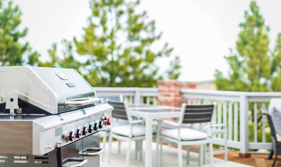 Een zilveren barbecue staat op een terras met een witte tafel en stoelen, tegen een achtergrond van groene bomen.