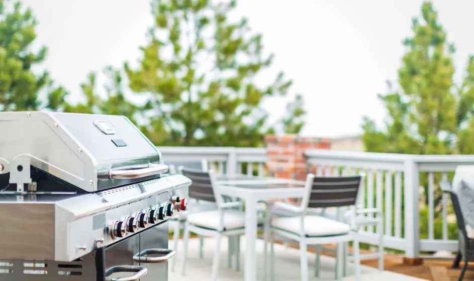 Een zilveren barbecue staat op een terras met een witte tafel en stoelen, tegen een achtergrond van groene bomen.