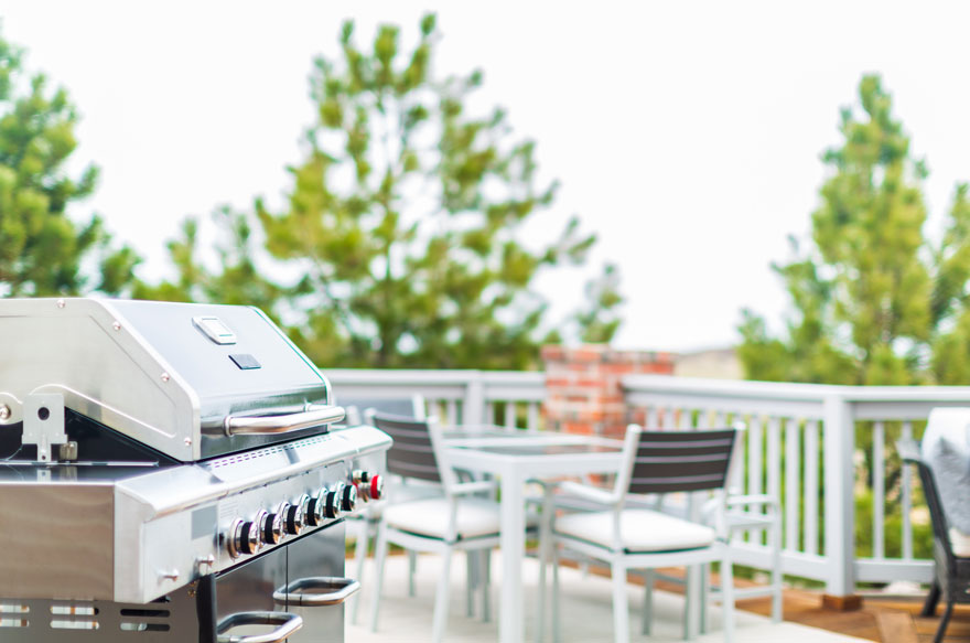 Un barbecue en acier inoxydable se trouve sur une terrasse avec une table et des chaises blanches. Des arbres sont visibles à l'arrière-plan.