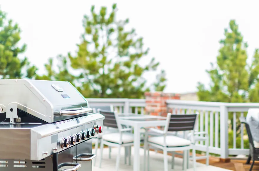 Un barbecue en acier inoxydable se trouve sur une terrasse avec une table et des chaises blanches. Des arbres sont visibles à l'arrière-plan.