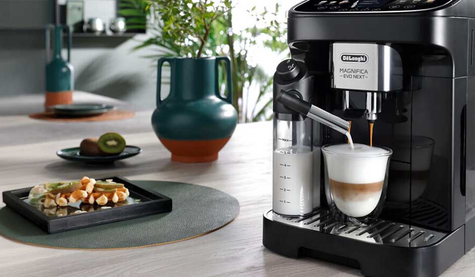 Une De'Longhi Magnifica prépare un cappuccino. Sur la table, un plateau de snacks, un kiwi et un vase décoratif.