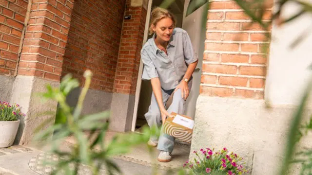 Une personne sort d'une porte en briques, tenant un colis. Des plantes sont de part et d'autre de l'entrée.