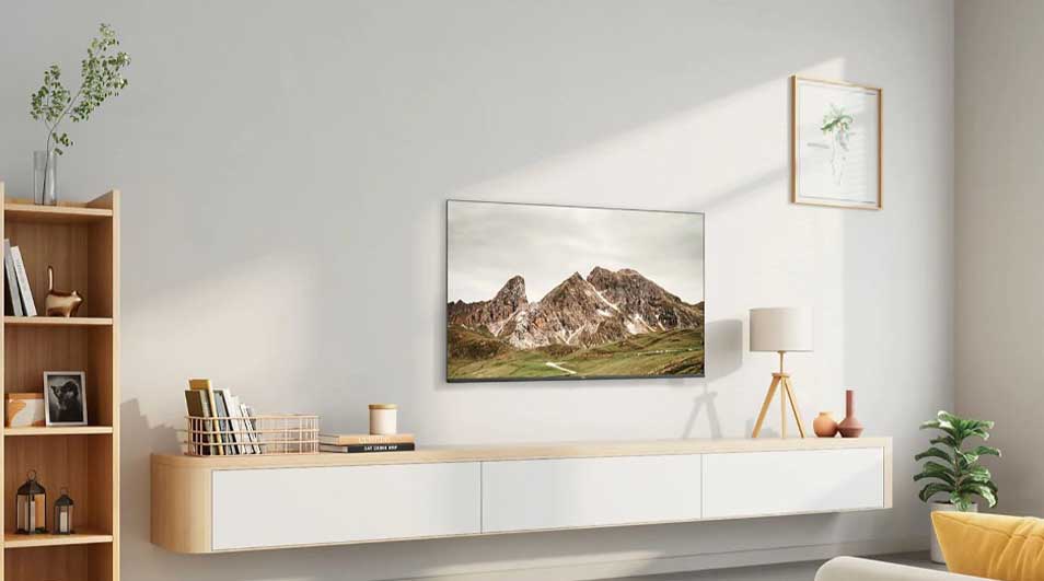 Salon lumineux avec TV montrant des montagnes, étagère en bois, et mobilier blanc. Une plante verte ajoute une touche de couleur.
