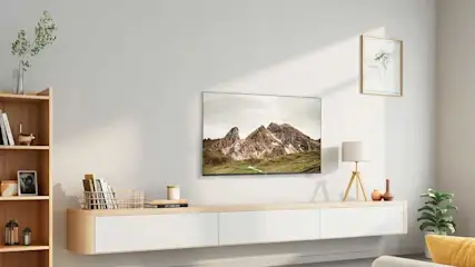 Woonkamer met tv met berglandschap, houten kast, lamp en groene planten tegen een witte muur.
