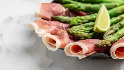 Een bord met groene asperges, opgerolde ham en een partje citroen op een witte achtergrond.