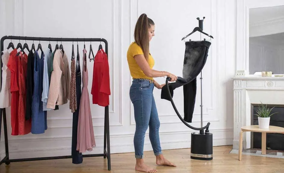 Een kledingstomer stoomt een zwarte broek. Een rek met kleding staat links. Een witte muur is op de achtergrond.
