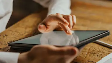 Handen bedienen een tablet op een houten tafel. Een zilveren stylus en een laptop zijn zichtbaar op de achtergrond.