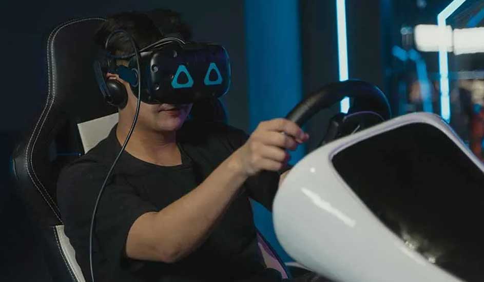 Un casque VR noir avec des triangles bleus est porté. On voit un volant noir et un simulateur blanc dans un environnement bleu.