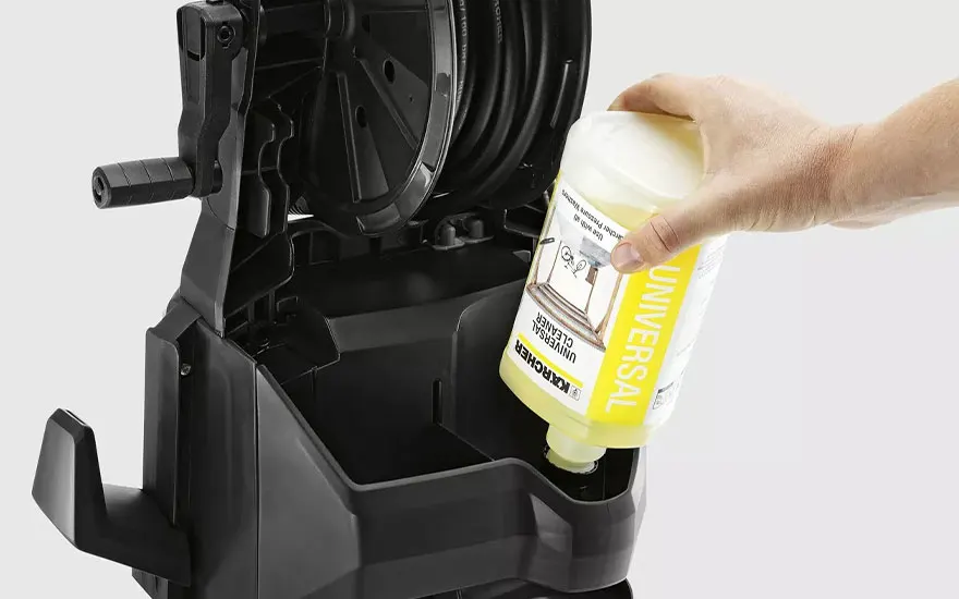 Een hand giet gele universele reiniger uit een fles in het reservoir van een zwarte hogedrukreiniger met opgerolde slang op een witte achtergrond.