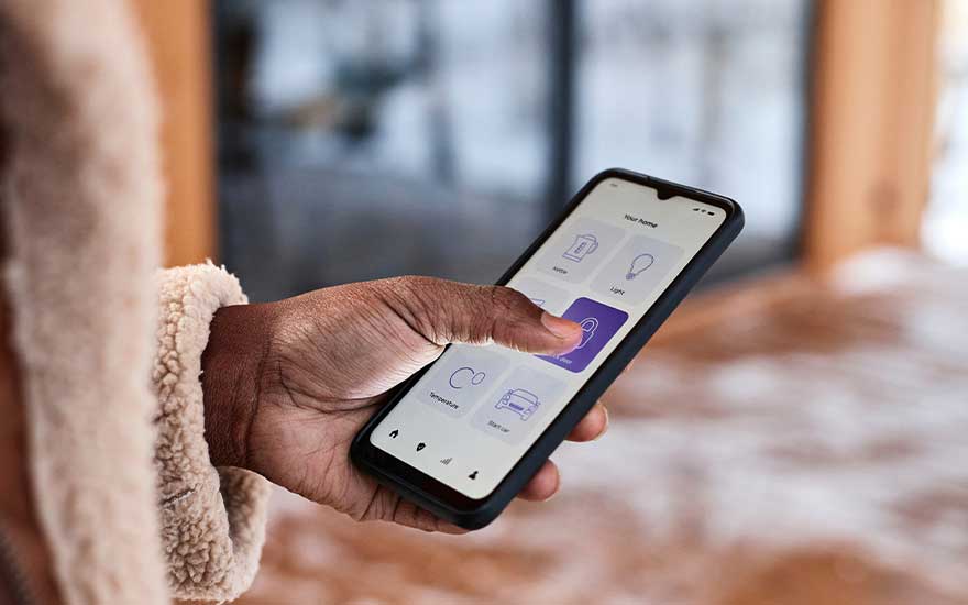 Een hand gebruikt een smartphone met smart home-bediening, waarop pictogrammen voor licht en apparaten zichtbaar zijn in huis.