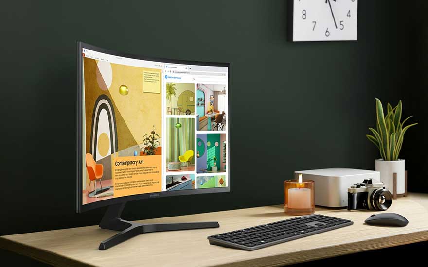Een gebogen monitor toont een website over kunst. Een toetsenbord, muis, camera en kaars staan op een houten bureau. Een klok hangt aan de groene muur.
