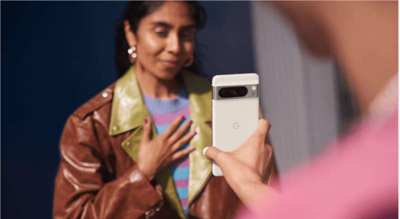 Une main tient un smartphone blanc avec un bloc caméra gris. Une veste en cuir marron est visible en arrière-plan.