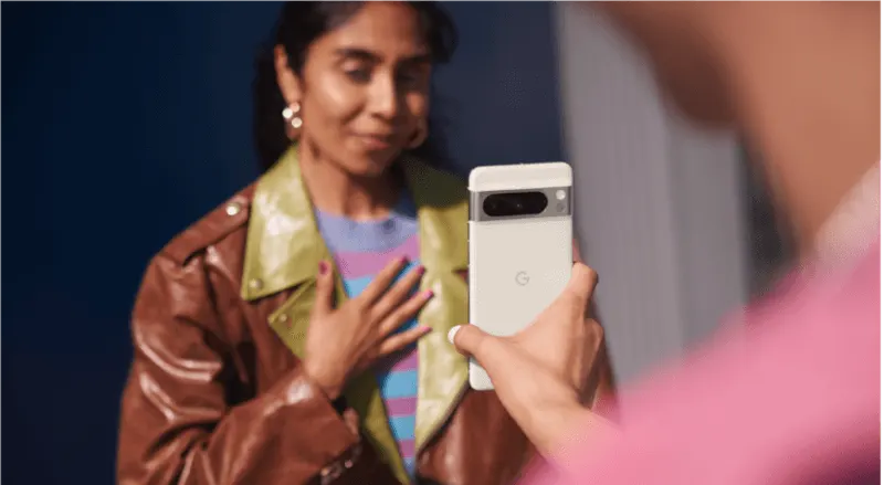 Une main tient un smartphone blanc avec un bloc caméra gris. Une veste en cuir marron est visible en arrière-plan.