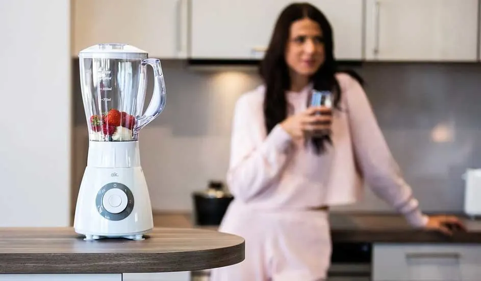 Een witte blender met aardbeien en bananen staat op een houten aanrecht in een keuken.