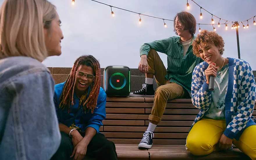 A group sits on a bench with a black speaker. String lights hang above. The speaker has a colorful, circular light display.
