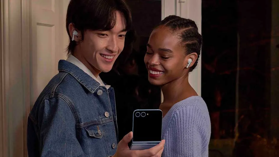 Un homme et une femme sourient en regardant un téléphone pliable argenté sur fond de porte blanche et de mur sombre.