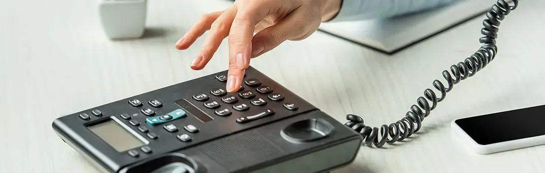 Een zwarte telefoon met knoppen en een snoer op een wit bureau. Een hand toetst een nummer in.