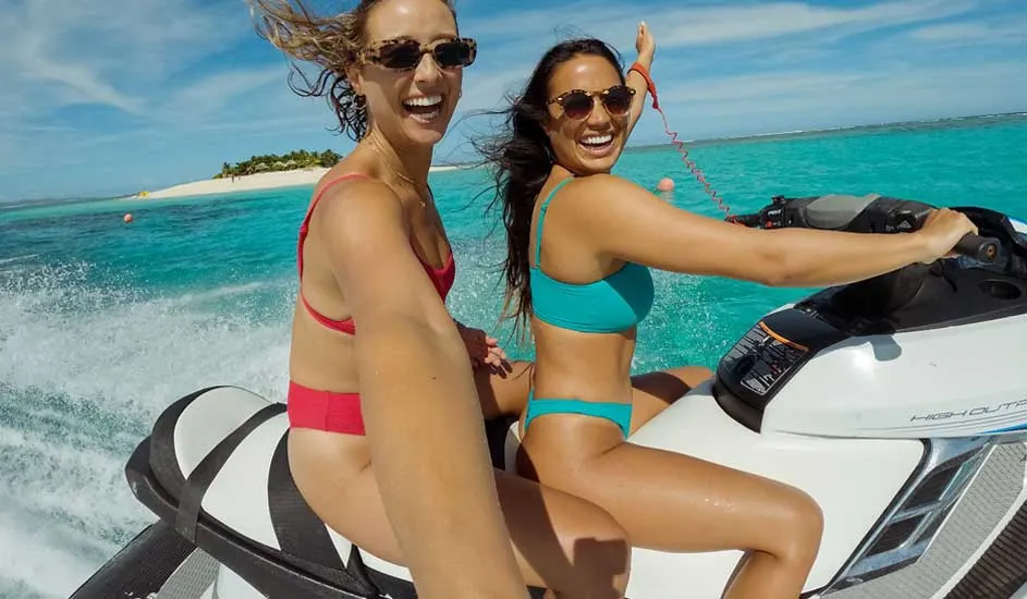 Twee vrouwen op een jetski in helderblauw water met een eiland op de achtergrond onder een blauwe hemel.