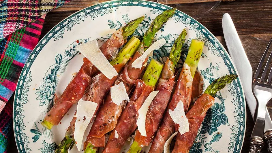 Asperges met ham en kaasschilfers op een bord met bloemmotief, naast een mes en vork.