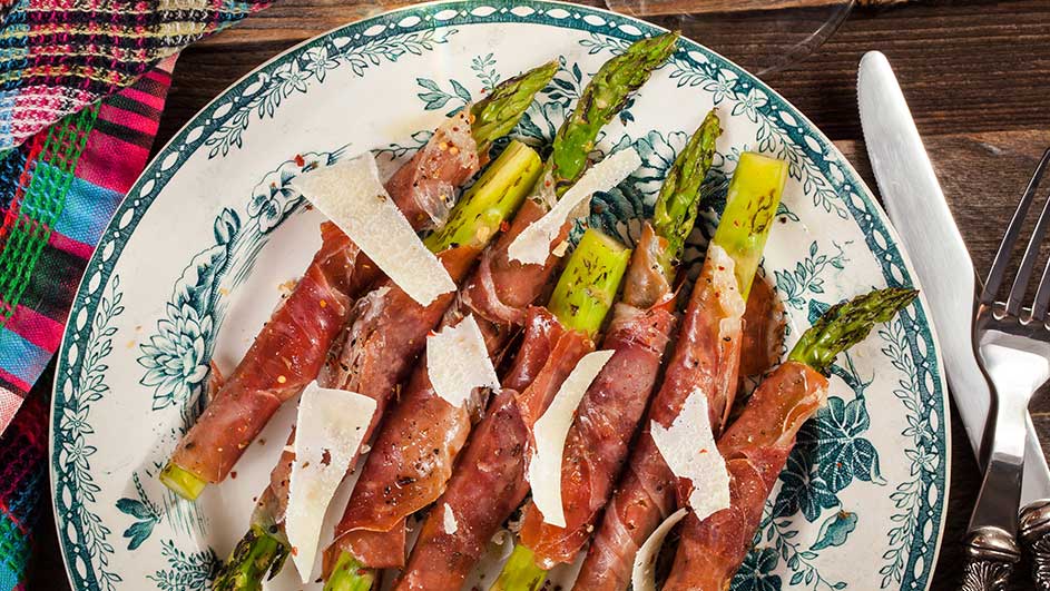 Assiette avec asperges vertes enroulées de jambon, copeaux de fromage, sur table en bois. Couteau et fourchette à droite.